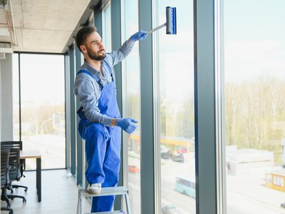 Gebäudereiniger reinigt Glasfassade von innen mit Fensterwischer und steht auf einer Leiter. Klicke auf das Bild, um auf die Seite Fensterreinigung zu kommen.
