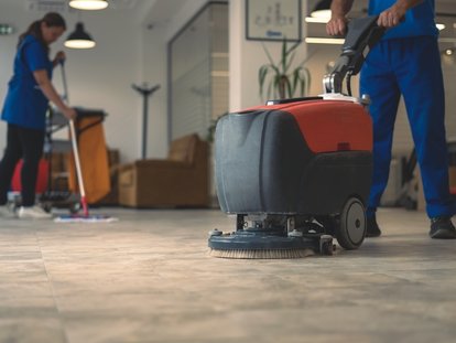 Reinigungskraft reinigt Boden mit einer Bodenreinigungsmaschine in Büro. Im Hintergrund putzt eine zweite Person mit einem Wischer den Boden. Klicke auf das Bild, um auf die Seite Grundreinigung zu kommen.